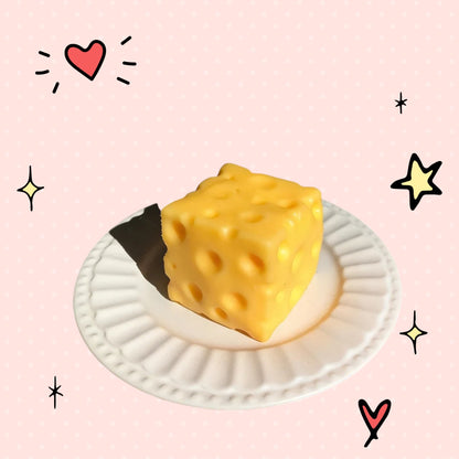 Foundry FitFit yellow cheese block squishy fidget toy on a small white plate with a playful pink background